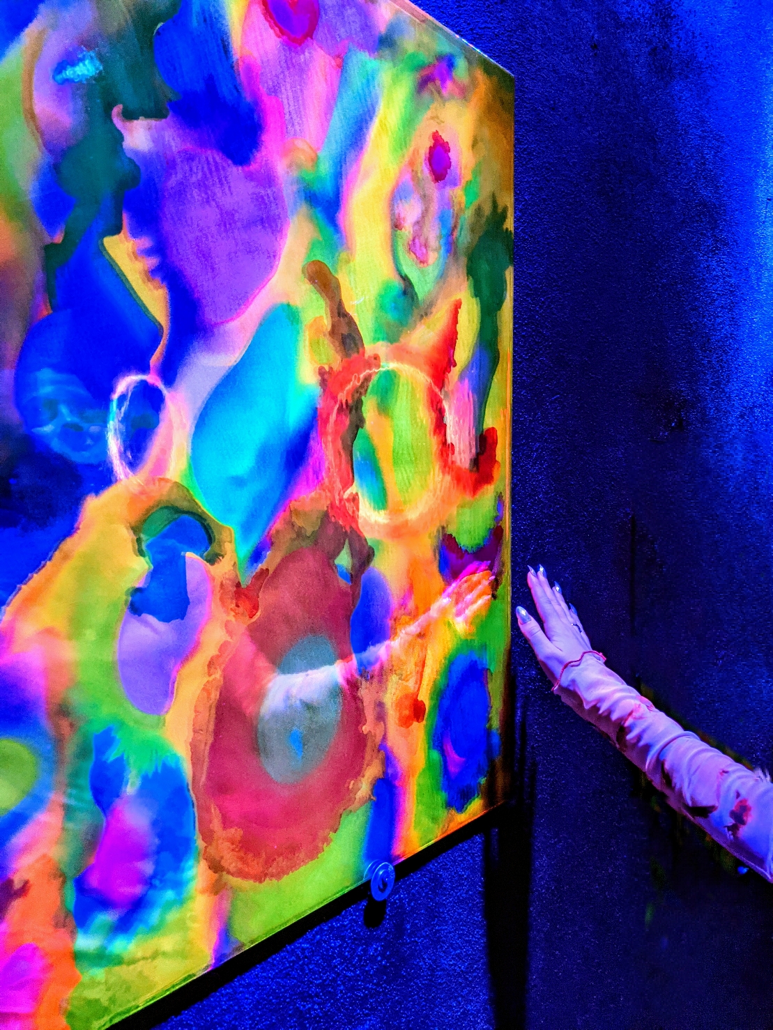 Large UV-reactive painting by Haiiileen installed in a corner space illuminated by deep blue lighting.