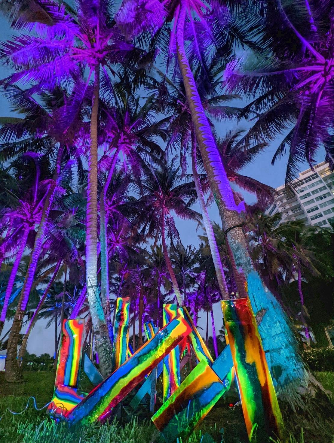Neon rainbow-painted wooden sculptures glowing beneath violet-lit palm trees at North Beach Music Festival.