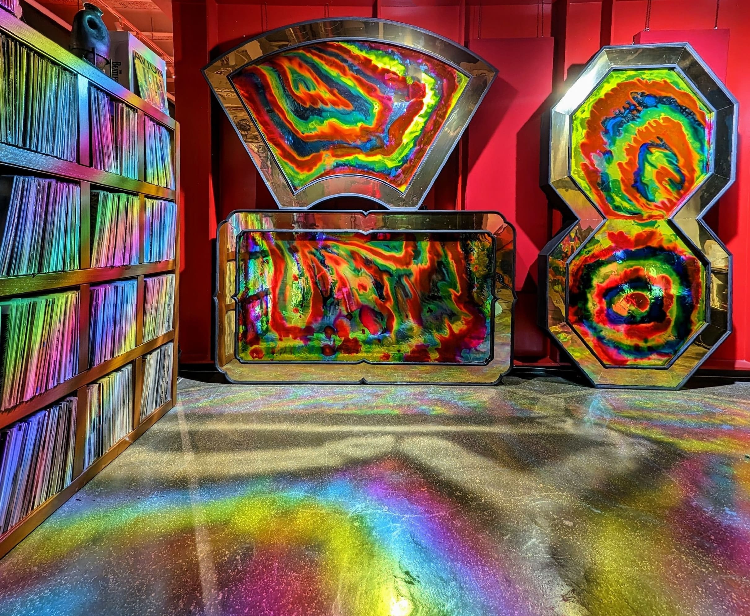 Neo-acid reflective mirror installation by Haiiileen for AVIÓN Tequila, casting rainbow light waves across vinyl shelves and the room.