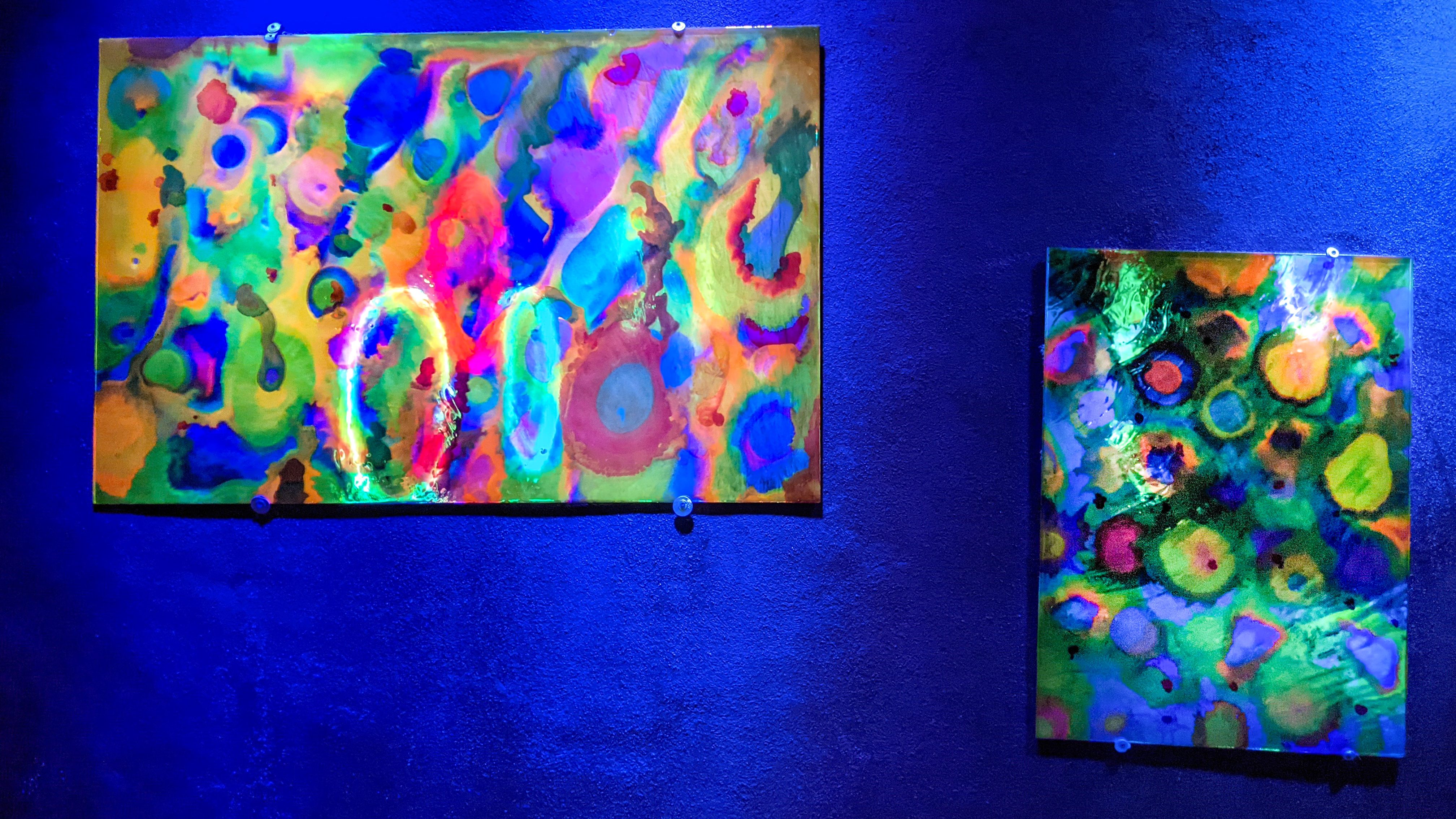 Large UV-reactive painting by Haiiileen installed in a corner space illuminated by deep blue lighting.