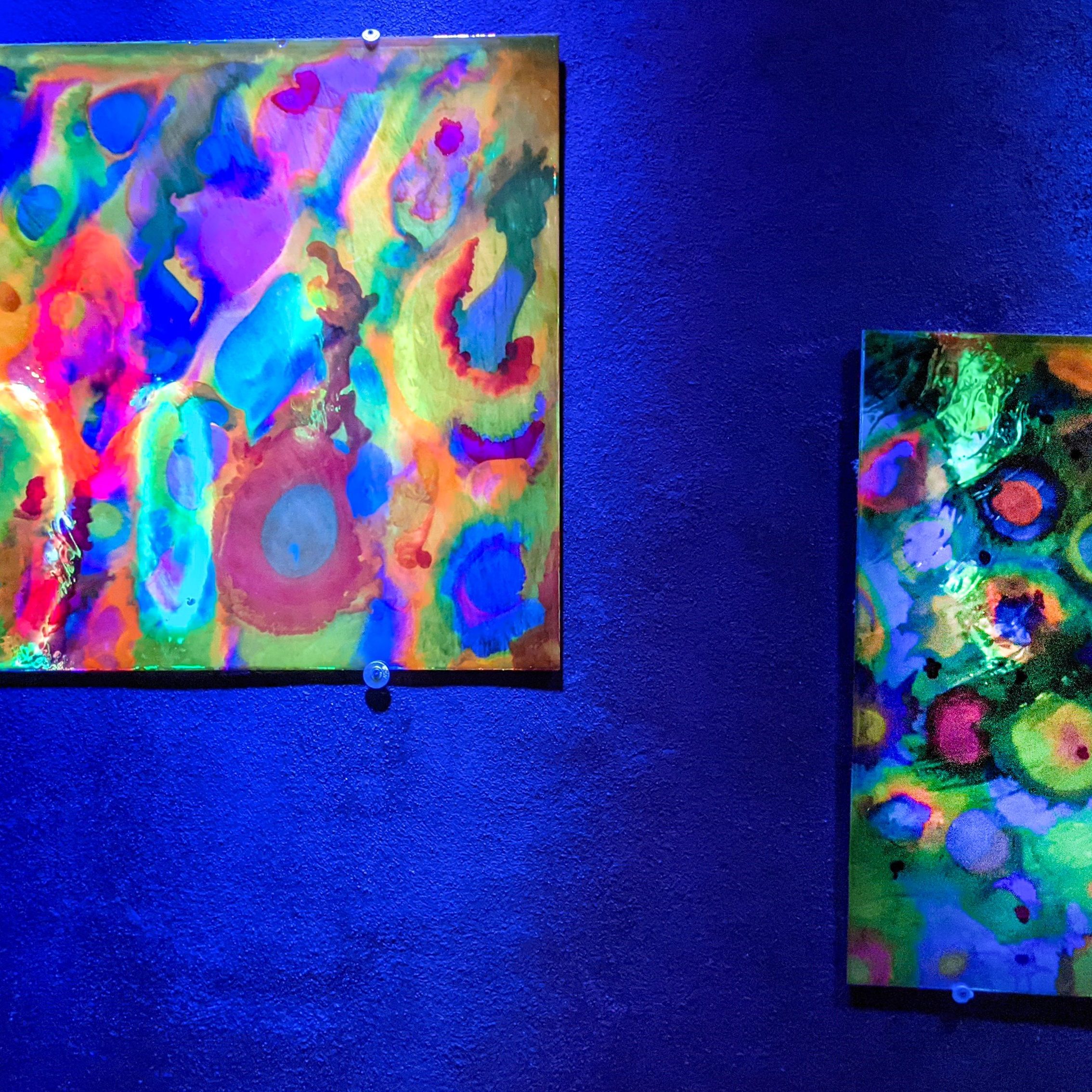 Large UV-reactive painting by Haiiileen installed in a corner space illuminated by deep blue lighting.